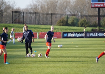 Individuals are engaged in a soccer training session on a grassy field. Some are dribbling soccer balls while a coach oversees the activities. In the background, there is a fence and a sign with the words 'Wash Spirit'.