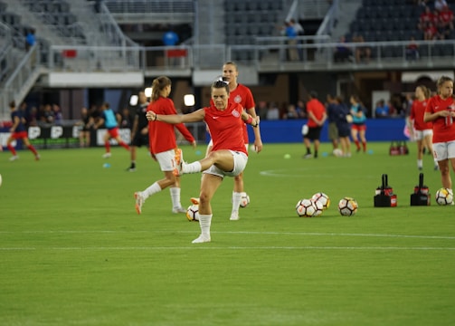 A group of soccer players in red jerseys and white shorts are practicing on a green field. One player in the foreground is stretching their leg, while others are engaged in warming up or training exercises. There are soccer balls scattered on the ground, and the setting appears to be a large sports stadium with empty stands in the background.