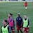 A group of female soccer players wearing training vests and sports attire walking on a green soccer field. Some players are wearing pink vests while others are in neon green. They are participating in a training session, with one player slightly in the background. The field is large and appears well-maintained, bordered in the distance by advertising boards.