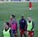 A group of female soccer players wearing training vests and sports attire walking on a green soccer field. Some players are wearing pink vests while others are in neon green. They are participating in a training session, with one player slightly in the background. The field is large and appears well-maintained, bordered in the distance by advertising boards.