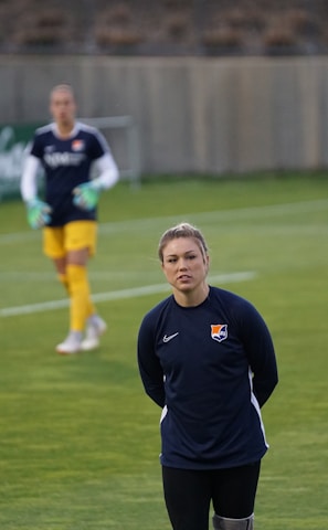 A person stands on a soccer field, wearing a navy sports jacket with a logo. In the background, another person in sports gear, including yellow shorts, is visible, holding goalkeeper gloves. A third person is near some soccer balls, facing away. The setting appears to be a practice session with advertising boards visible along the sidelines.