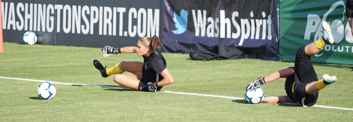 Two football players are performing a training drill on a grassy field. One is leaning back while controlling a soccer ball with her hands, and the other is lying on her back with her legs and arms up, holding another soccer ball. Both are wearing black attire with yellow socks. In the background, banners display social media handles and web addresses, and a person in a blue shirt stands nearby, watching the scene.