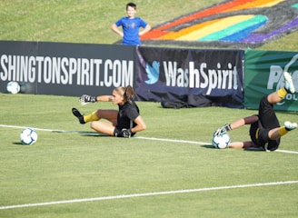 Two football players are performing a training drill on a grassy field. One is leaning back while controlling a soccer ball with her hands, and the other is lying on her back with her legs and arms up, holding another soccer ball. Both are wearing black attire with yellow socks. In the background, banners display social media handles and web addresses, and a person in a blue shirt stands nearby, watching the scene.