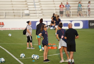 A group of people are gathered on a soccer field, likely during a training session. Some individuals are wearing athletic gear and appear to be coaching or instructing, while others are moving in the background. There are soccer balls scattered on the grass, and orange cones mark certain areas. A nearby bench is visible with some seated spectators.