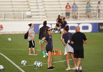 A group of people are gathered on a soccer field, likely during a training session. Some individuals are wearing athletic gear and appear to be coaching or instructing, while others are moving in the background. There are soccer balls scattered on the grass, and orange cones mark certain areas. A nearby bench is visible with some seated spectators.