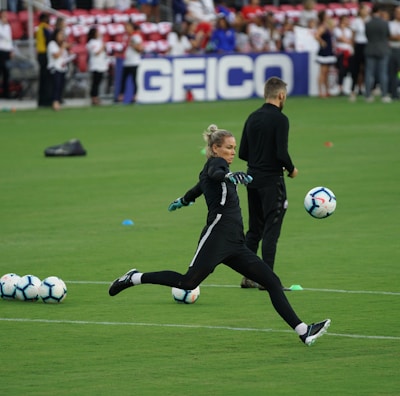 A soccer player in a black sports outfit is mid-action on a green field, focusing intently on kicking a soccer ball. Several soccer balls are scattered on the grass nearby, and a coach or teammate stands in the background watching. Spectators can be seen in the distant stands.