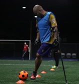 A man wearing a blue training vest is practicing on a football field. He is using crutches and moving a bright orange ball with his foot. Other people in sports attire can be seen in the background, and the field is marked with orange cones.