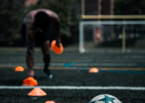 A person is engaged in a soccer training session on an outdoor field. The individual is focused on maneuvering through orange cones arranged on the grass. In the foreground, a soccer ball rests on the ground. There are buildings and trees in the background under a partly cloudy sky.