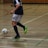 A person in a navy blue and white sports uniform is playing futsal in an indoor court. The individual is about to kick a colorful ball while wearing bright green and black shoes. The court is marked with various colored lines and has a wooden wall in the background.